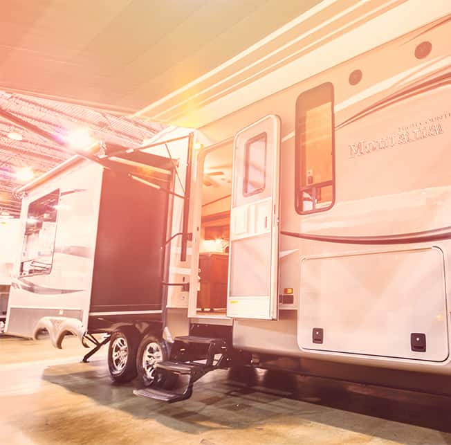 A spacious RV with open slide-outs is parked indoors, exuding a home-like vibe. The door, slightly ajar, offers a peek into its inviting interior. Set in a well-lit showroom or exhibition space, the scene highlights the RV's modern design and homely charm.