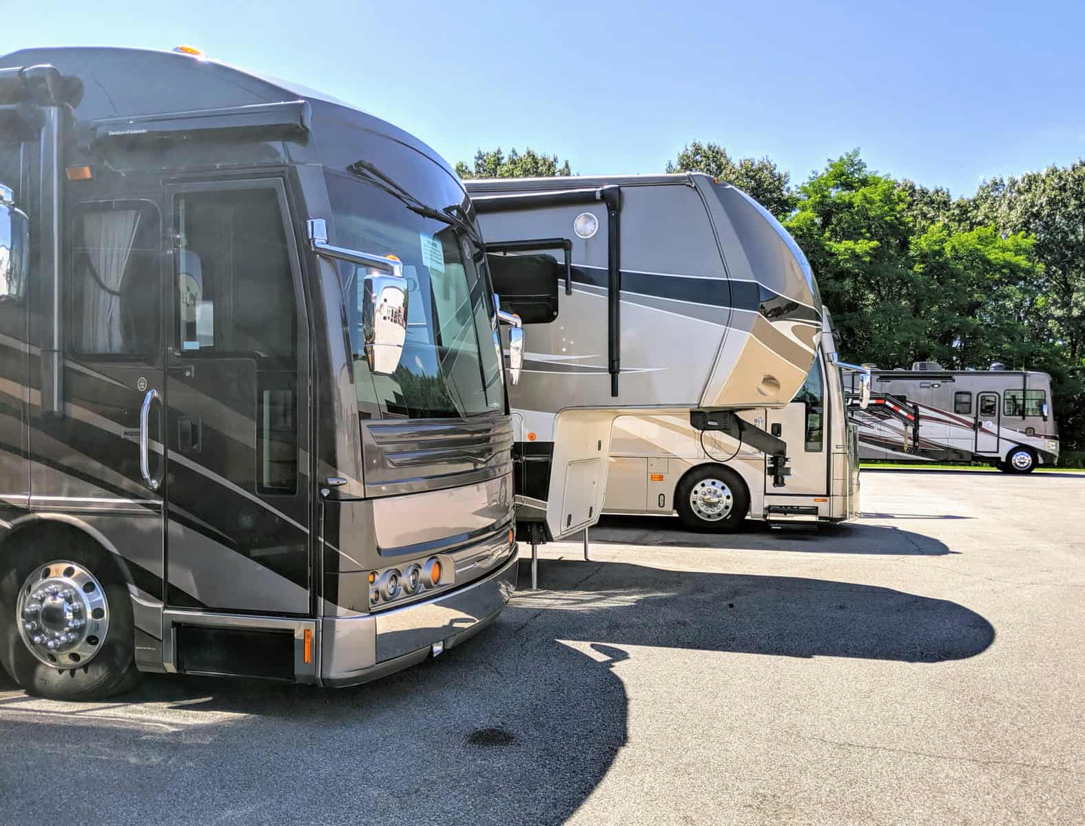 A row of recreational vehicles (RVs) parked in a lot under a clear blue sky. The foreground shows two large RVs, offering home-like comforts up close, while another RV is nestled in the background, surrounded by the tranquility of trees.
