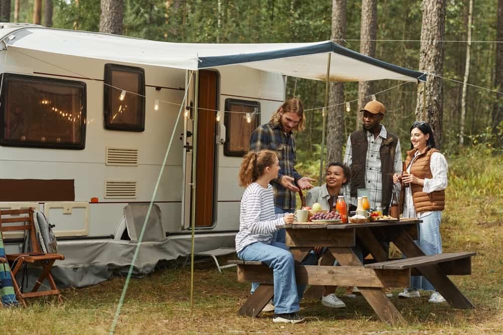 Five people enjoy a meal at a picnic table next to a camper in a forest. The table has food and drinks, and string lights adorn the camper. Everyone is smiling and engaged in conversation.
