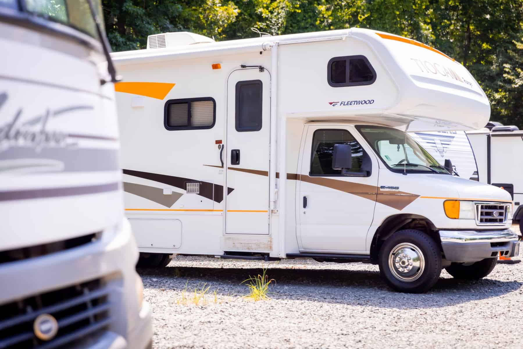 A parked white and orange Fleetwood RV, resembling a cozy home on wheels, is seen in a sunlit gravel lot with trees in the background. Another vehicle is partially visible on the left side of the image.
