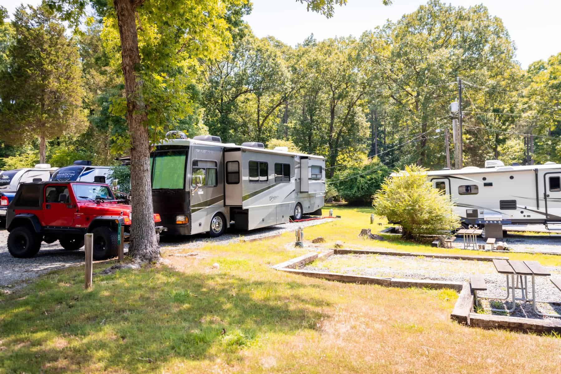 A campground feels like home with several RVs parked on gravel spots surrounded by trees. A red Jeep rests beside a large grey RV, creating a cozy retreat. A picnic table and fire pit sit in the foreground, offering a gathering space shaded by grass and trees.
