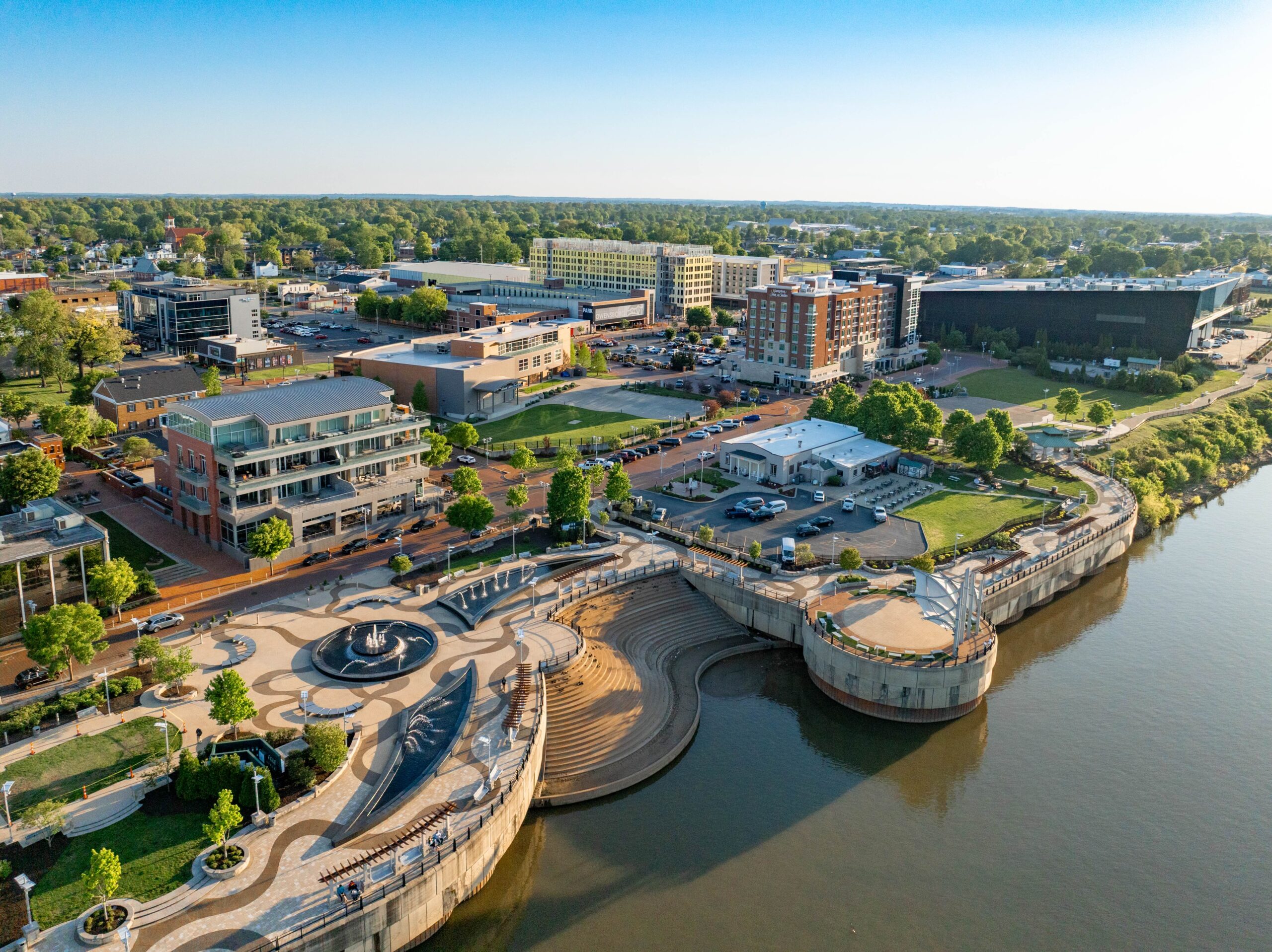 Aerial view of a riverside park with curved walkways, steps, fountains, and nearby buildings under a clear sky.